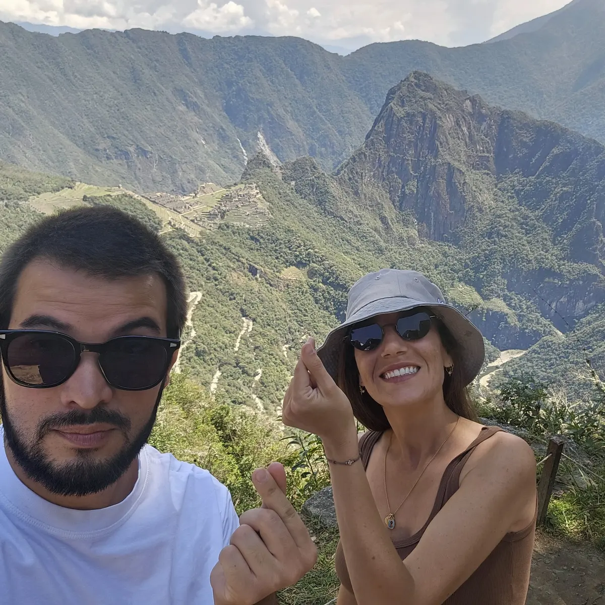 Holiday selfie of a couple at Machu Picchu, both in sunglasses, making finger hearts